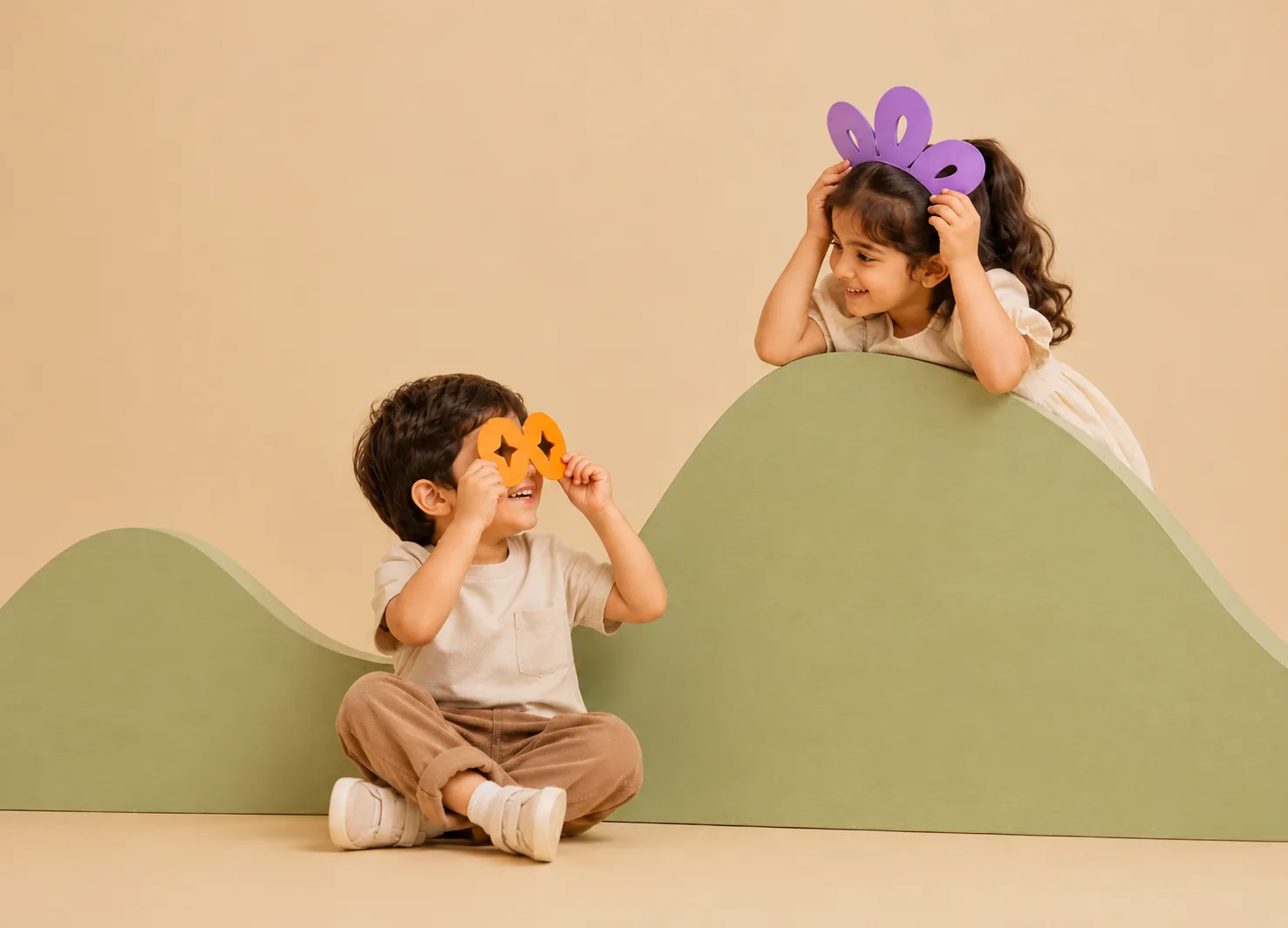 Two children playing on soft green hills, one wearing a purple paper crown